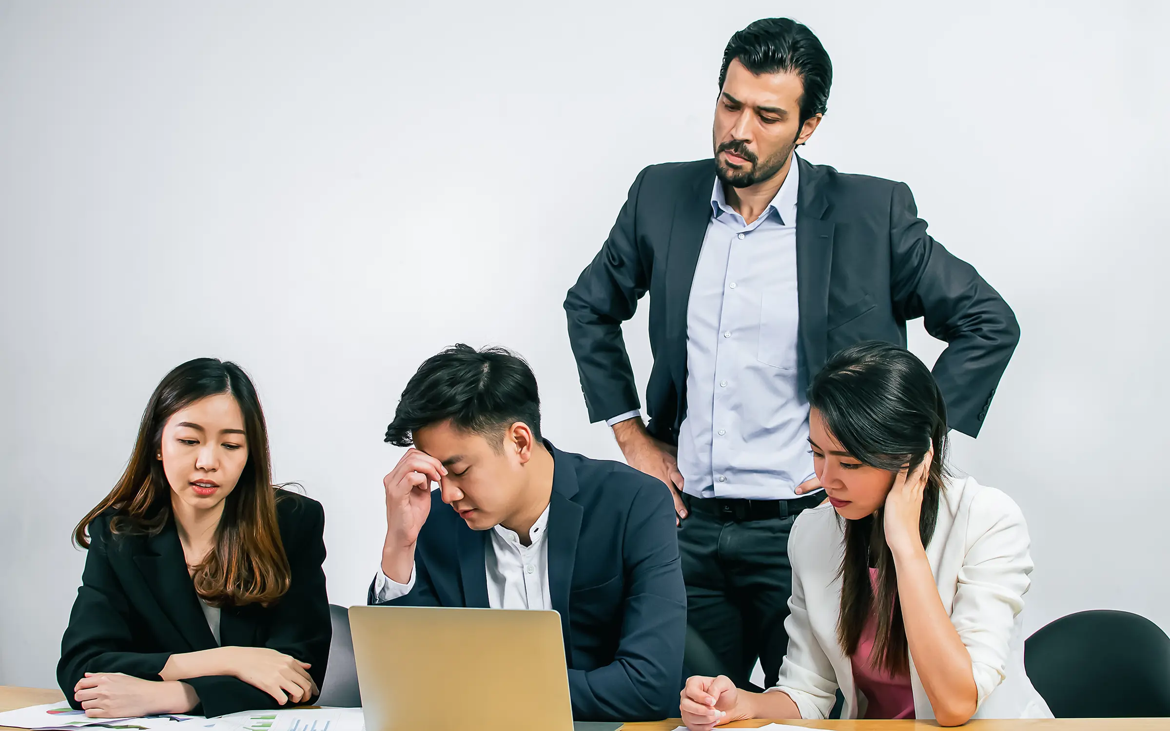 Four colleagues around a desk in a tense meeting: three seated and looking down, one standing over them with hands on hips.
